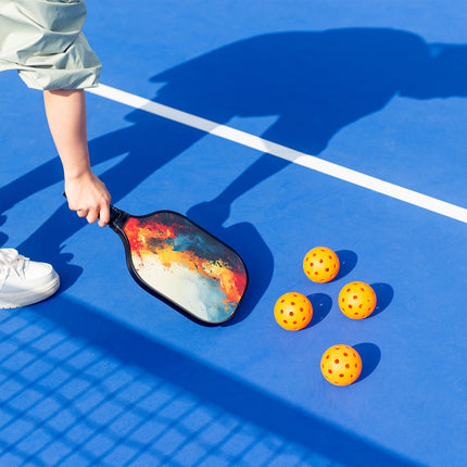 Person holding a colorful paddle with pickleball balls on a blue court
